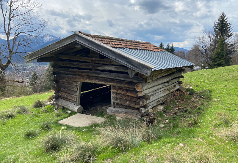 Holzhütte auf einer Wiese mit Bergen im Hintergrund.