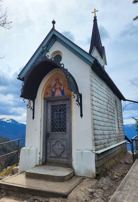 Kapelle mit Holzschindeln und Berglandschaft im Hintergrund.