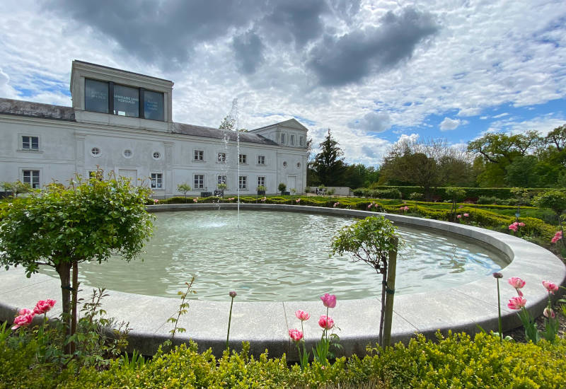 Brunnen in einem Garten mit Blumen und historischem Gebäude.