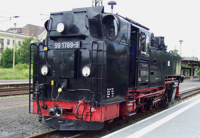 Schwarze Dampflok steht am Bahnhof, Fahrer schaut hinaus.
