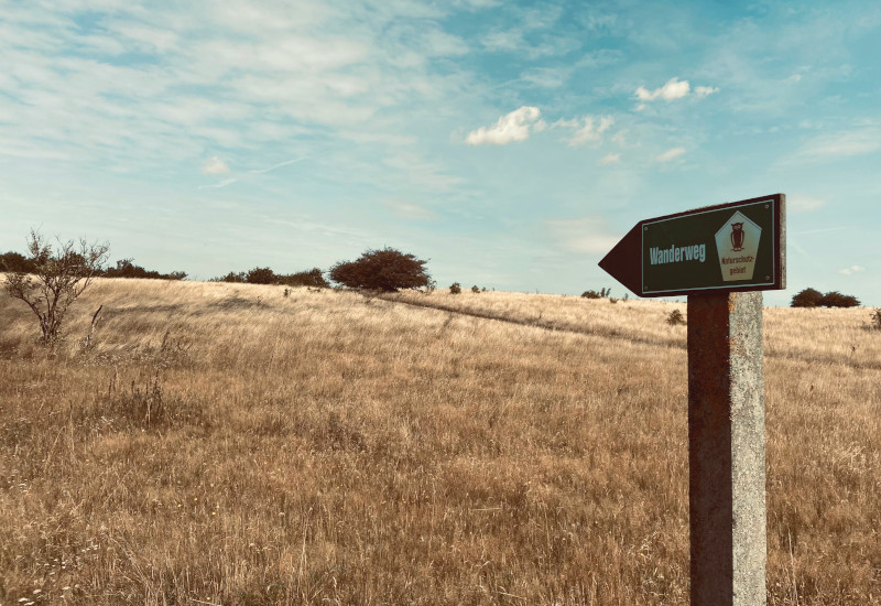 Wegweiser zum Wanderweg in einer hügeligen Landschaft.