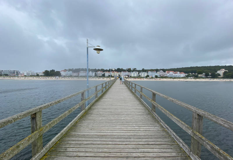 Holzsteg führt über Wasser zu einem Strand mit Häusern.