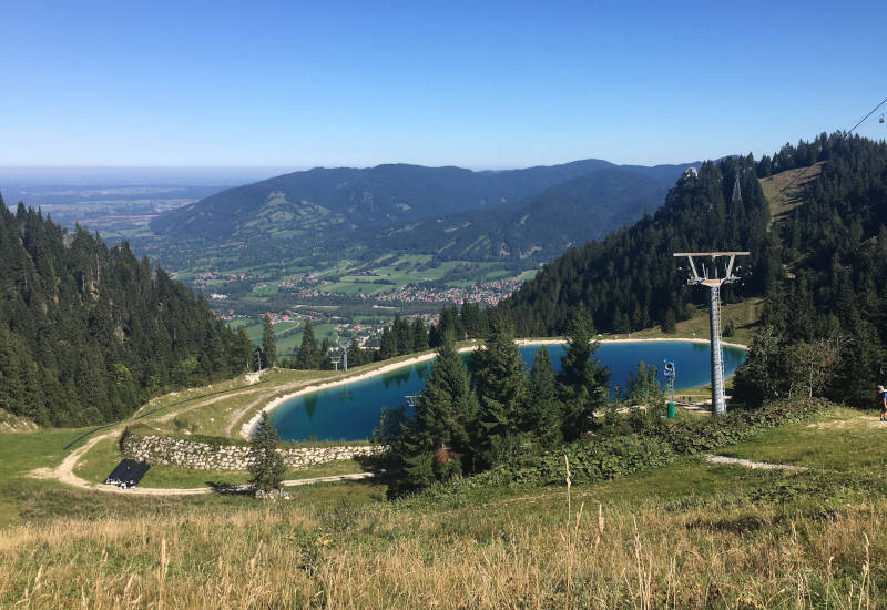 Berglandschaft mit See, Wäldern und Seilbahn im Hintergrund.