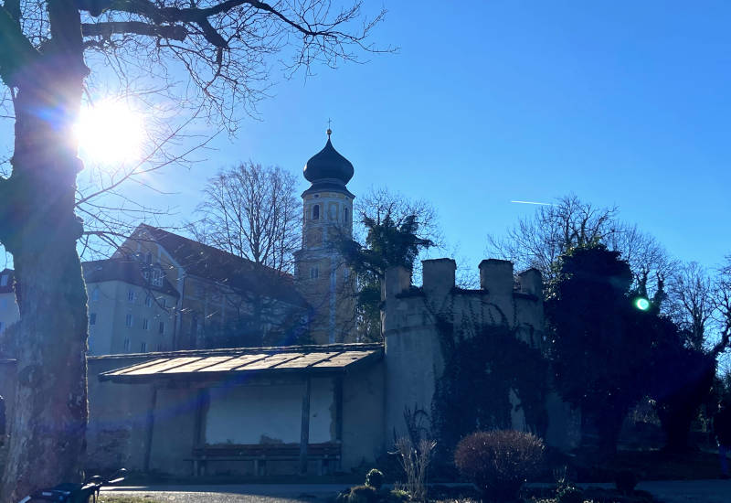 Kirchturm hinter Bäumen und Mauer, strahlender Sonnenschein.