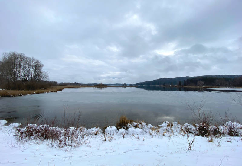 Winterliche Landschaft mit See, Schnee und bewölktem Himmel.