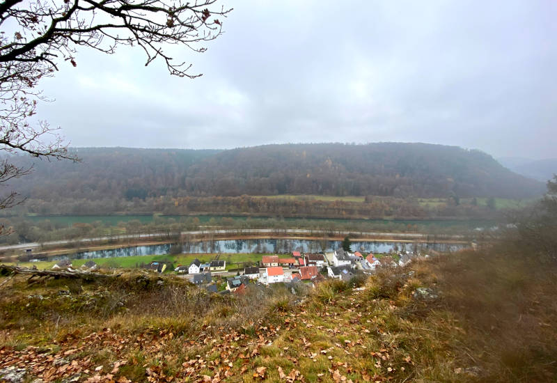 Blick auf ein Dorf am Fluss, umgeben von Hügeln.