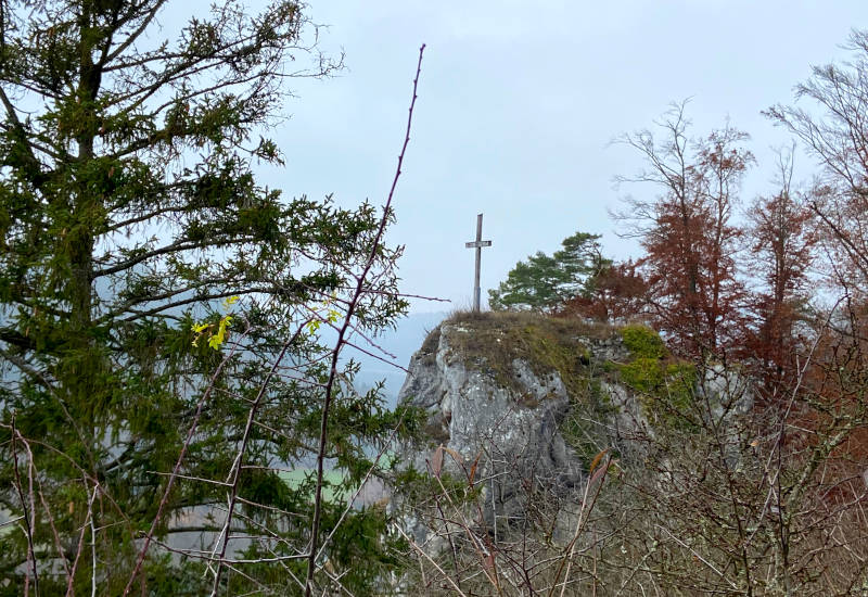 Kreuz auf einem Felsen, umgeben von Bäumen und Nebel.